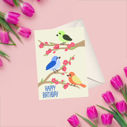 A playful birthday card design featuring three colorful birds perched on tree branches decorated with pink flowers. The soft and cheerful illustration is set on a clean background, making the artwork and colors pop. A whimsical and handmade card perfect for birthdays. coming with a matching white blank envelope