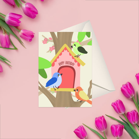 Greeting card featuring 3 colorful birds around a light pink birdhouse, the birdhouse hosts the text happy birthday above the door. Behind the card is a white blank envelope visable
