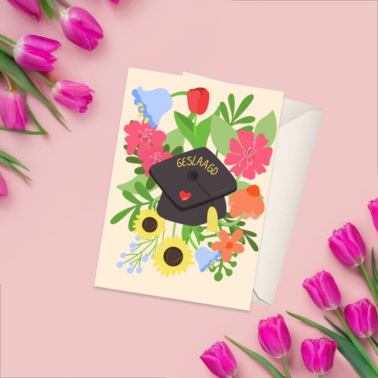 Greeting card on a beige background with a graduation cap with geslaagd on it. around the graduation cap there are vibrant flowers and greenery. On the back there is a matching white blank envelope