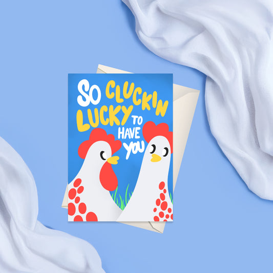 Bright blue card featuring two white chickens with red accents and a funky looking text saying I am so clucking lucky to have you. on the back of the card there is a matching white blank envelope visible 