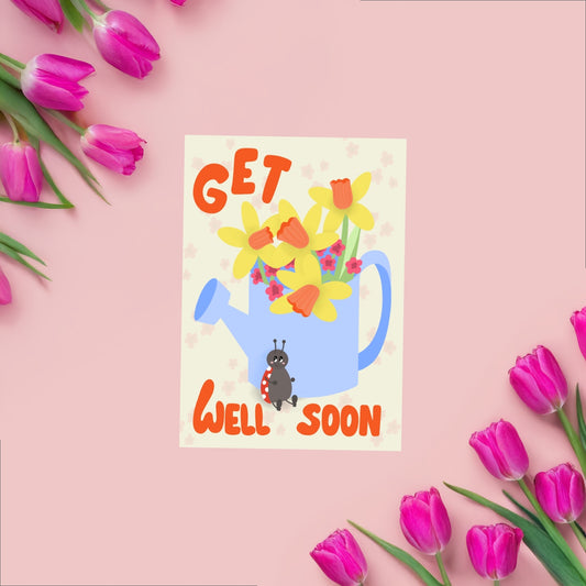 Greeting card wit a beige background featuring a blue watering can with yellow narcissus and other pink flowers comming out of it. on the bottom of the watering can is a ladybug sitting around the watering can there is the text get well soon in a bright orange color